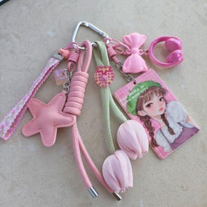 May include: A collection of pink keychains and charms, including a star, bow, headphones, and a card with a cartoon girl. The keychains are attached to a silver carabiner clip. The background is a light beige color.