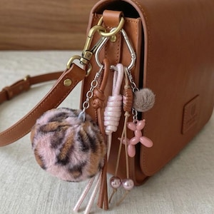May include: A tan leather crossbody bag with a gold-tone metal clasp featuring a collection of charms including a large pink and brown leopard print faux fur pom-pom, a pink knotted cord, a small brown pom-pom, and a pink balloon dog charm.