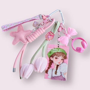 May include: A collection of pink keychains and charms. The keychains include a pink star, a pink bow, a pink headset, and a charm with a girl illustration. The keychains are attached to a silver carabiner clip.