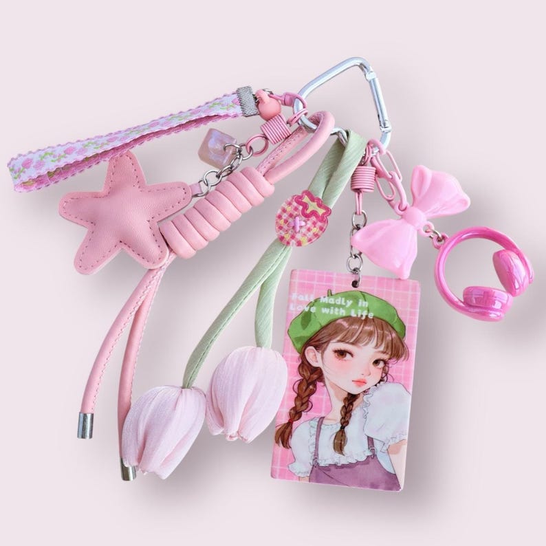 May include: A collection of pink keychains and charms. The keychains include a pink star, a pink bow, a pink headset, and a charm with a girl illustration. The keychains are attached to a silver carabiner clip.