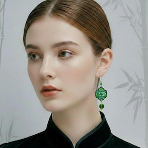 May include: A woman wearing green and blue drop earrings. The earrings feature a geometric pattern and a small green bead. The earrings are silver and are suspended from the woman's ear.