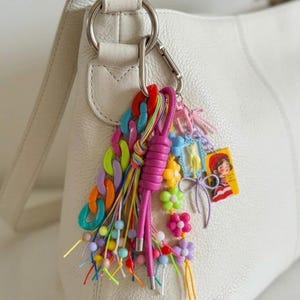 Puede incluir: Un colgante para bolso multicolor sujeto a un bolso de cuero blanco con un mosquet&oacute;n plateado. El accesorio incluye un cord&oacute;n anudado rosa brillante, una cadena de pl&aacute;stico gruesa en colores ne&oacute;n, cuentas pastel, dijes de flores y un colgante rectangular que muestra a una persona con una boina roja.