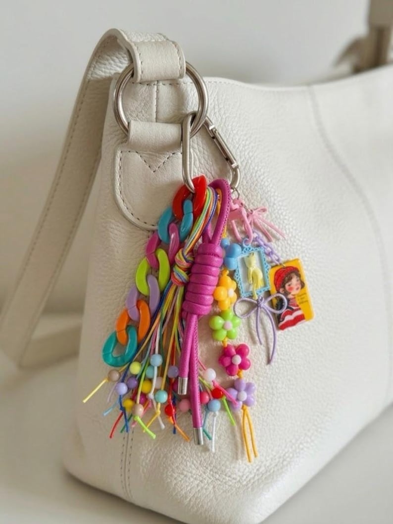 Puede incluir: Un colgante para bolso multicolor sujeto a un bolso de cuero blanco con un mosquet&oacute;n plateado. El accesorio incluye un cord&oacute;n anudado rosa brillante, una cadena de pl&aacute;stico gruesa en colores ne&oacute;n, cuentas pastel, dijes de flores y un colgante rectangular que muestra a una persona con una boina roja.