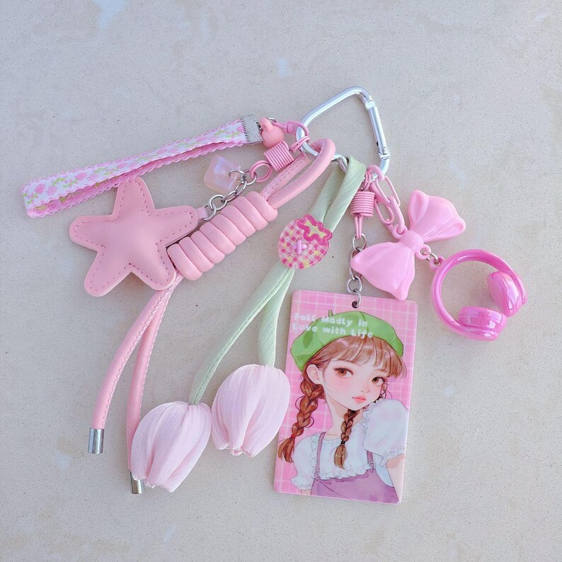 May include: A collection of pink keychains and charms. Includes a pink star, a bow, a pair of tulip-shaped charms, and a rectangular charm with a cartoon girl. The keychains are attached to a silver carabiner.