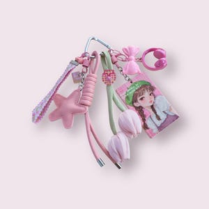 May include: A collection of pink keychains and charms, including a star, bow, headphones, and a card with a girl illustration. The keychains are attached to a silver carabiner clip. The color palette is primarily pink and pastel tones.