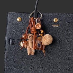 May include: A black leather tote bag with a gold metal logo that says "BURBERRY LONDON ENGLAND". The bag has a brown leather strap and a charm attached to it. The charm features a brown teddy bear, a small brown bear keychain, a brown pom-pom, and a few other decorative charms.