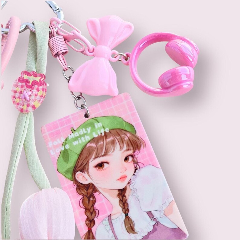 May include: A pink keychain with multiple charms. The charms include a pink bow, a pink headset, and a rectangular tag with an illustration of a girl wearing a green beret. The tag reads "Fall Madly in Love with Life".