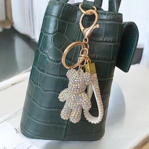 Op de afbeelding: Een goudkleurige sleutelhanger met een sprankelende, met strass bezaaide teddybeer-bedel en een witte polsband. De sleutelhanger is bevestigd aan een donkergroene, getextureerde handtas.