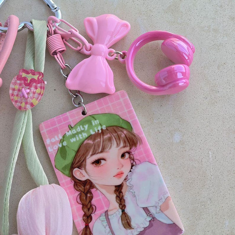 May include: A pink keychain with various charms, including a bow, headphones, and a tag with a cartoon girl wearing a green beret. The tag reads "Fall Madly in Love with Life."
