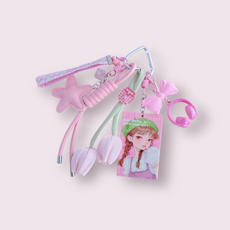 May include: A pink keychain with multiple charms, including a star, tulips, a bow, and headphones. The keychain also features a card with a cartoon girl and the text "sweetly in love with life".