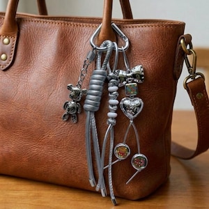 May include: A brown leather handbag with a silver-toned metal carabiner attached to the handle. The carabiner holds several charms, including a metallic teddy bear, a silver cord knot, white beads, a bow, a heart charm, and circular pendants.