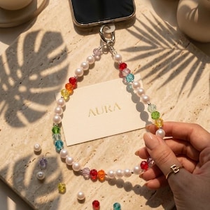 May include: A phone charm strap featuring a repeating pattern of white faux pearls and multi-colored faceted crystal beads in red, orange, yellow, green, blue, and purple. The strap attaches to a smartphone with a silver-toned metal ring.