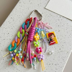 Puede incluir: Un llavero multicolor con un mosquet&oacute;n plateado, un cord&oacute;n de cuero rosa grueso, una cadena de pl&aacute;stico gruesa en naranja, morado, verde y azul, y varios dijes que incluyen un gato amarillo en un marco azul y una etiqueta rectangular con una ilustraci&oacute;n.