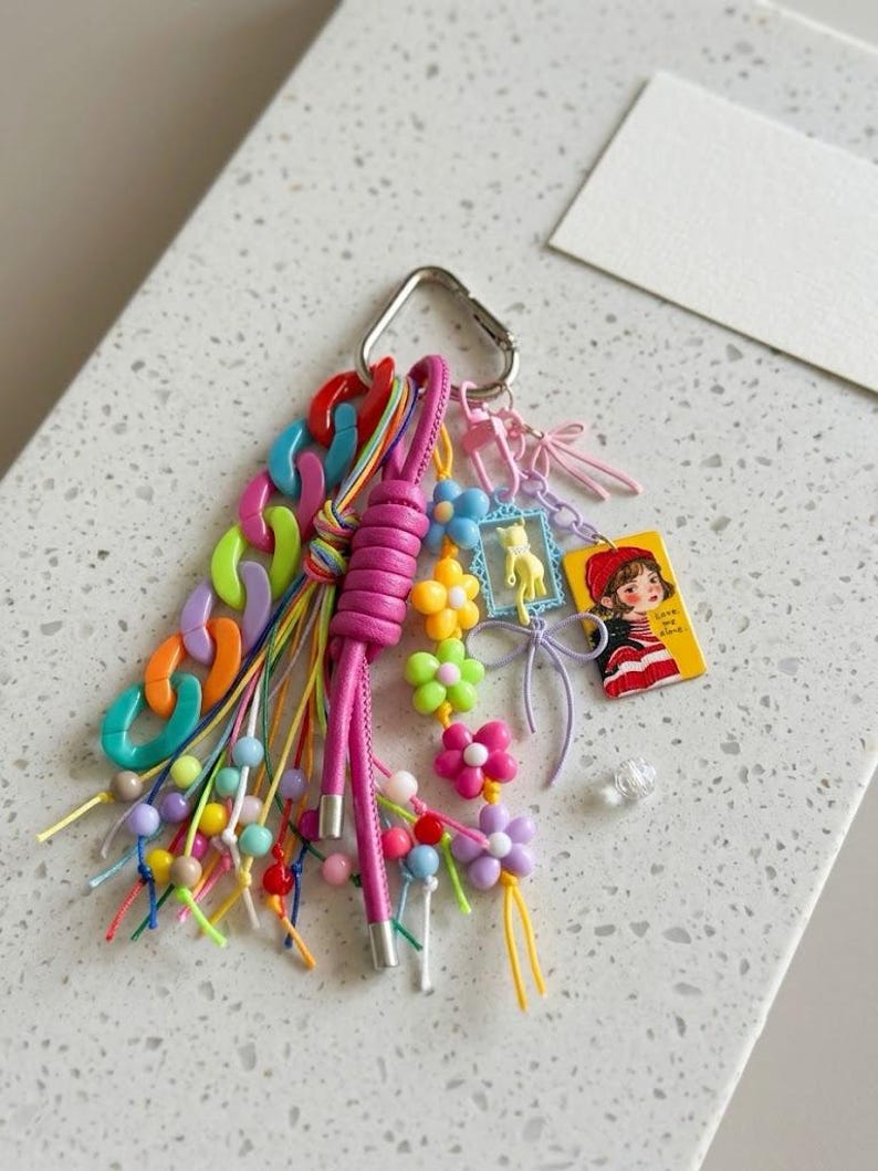 Puede incluir: Un llavero multicolor con un mosquet&oacute;n plateado, un cord&oacute;n de cuero rosa grueso, una cadena de pl&aacute;stico gruesa en naranja, morado, verde y azul, y varios dijes que incluyen un gato amarillo en un marco azul y una etiqueta rectangular con una ilustraci&oacute;n.