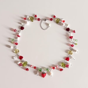 Puede incluir: Un delicado collar con un diseño caprichoso de fresa. El collar está adornado con cuentas rojas, verdes y blancas, junto con dijes en forma de hoja y corazón. El collar está hecho de metal color plata y tiene un cierre de cadena.