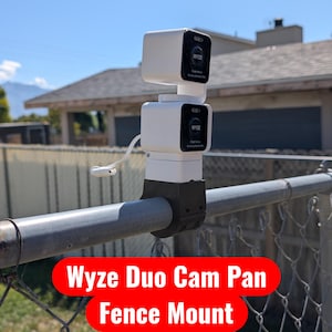 Peut inclure: Un support de caméra Wyze Duo Cam Pan blanc est fixé à une clôture métallique. Le support comporte deux unités de caméra carrées empilées avec le logo Wyze. Le support est noir et blanc, avec un câble sortant de l'unité supérieure. Le texte "Wyze Duo Cam Pan Fence Mount" est dans une bannière rouge.
