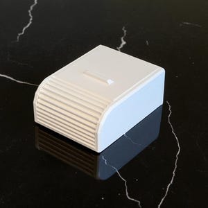 May include: A small, rectangular, white box with a light blue base. The front of the box has a series of horizontal, ridged lines. The top of the box has a small, rectangular indentation. The box is sitting on a black surface.