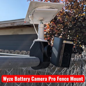 Peut inclure: Support de caméra de batterie Wyze Battery Camera Pro noir avec un panneau solaire blanc et une clôture argentée. La caméra a une lumière intégrée et le texte "WYZE" est visible. Le texte "Wyze Battery Camera Pro Fence Mount" est dans une bannière rouge.