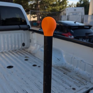 May include: A black metal truck bed stake with an orange plastic cap. The stake is in the bed of a white pickup truck.