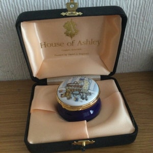 May include: A blue and gold enamel trinket box with a painted scene of a baby in a crib with a teddy bear. The box is in a black velvet box with the words "House of Ashley, Copper Enamels, Painted by Hand in England" on the inside.