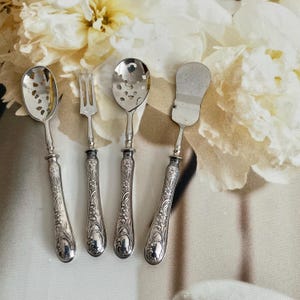 May include: A set of four antique silver serving utensils with floral patterned handles. The set includes a slotted spoon, a fork, a serving spoon, and a spatula-like knife. The utensils are displayed against a backdrop of white and cream-colored flowers.