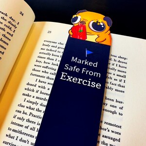 May include: A navy blue bookmark with the text "Marked Safe From Exercise" and a cartoon pug holding a juice box. The bookmark is inserted into a book with printed text.