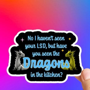 May include: A sticker with a black background and white border. It features two cartoon dragons, one white and one green, with the text "No I haven't seen your LSD, but have you seen the Dragons in the kitchen?"