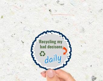 May include: A white sticker with a blue border and a white background. The sticker has a green recycling symbol, an orange arrow, and the text "Recycling my bad decisions daily".
