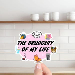 May include: A sticker with the text "THE DRUDGERY OF MY LIFE" in black, surrounded by cartoon images of chores and a corgi. The sticker is pink and is held up by a hand.