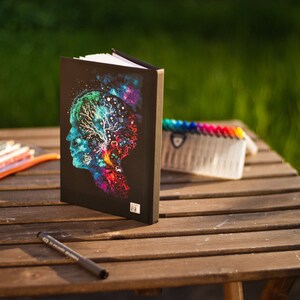 May include: A black journal with a colorful illustration of a human profile with a tree-like design inside. A clear pencil case with colored pencils, a black pen, and a set of colorful markers are on a wooden table.