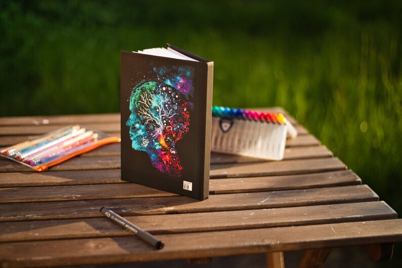 May include: A black journal with a colorful illustration of a human profile with a tree-like design inside. A clear pencil case with colored pencils, a black pen, and a set of colorful markers are on a wooden table.