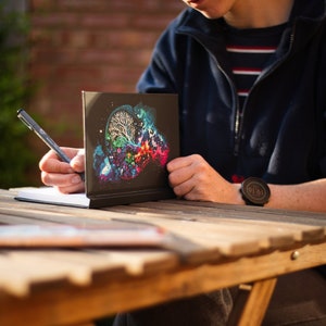 May include: A black notebook with a colorful brain-like design is propped up on a wooden table. A person is writing in a blank notebook with a pen. The scene is outdoors, with sunlight and greenery in the background.