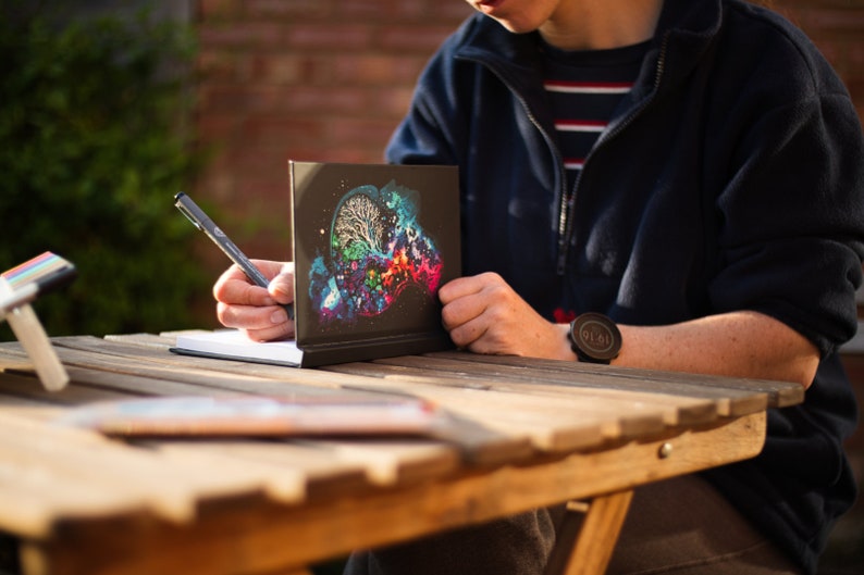 May include: A black notebook with a colorful brain-like design is propped up on a wooden table. A person is writing in a blank notebook with a pen. The scene is outdoors, with sunlight and greenery in the background.