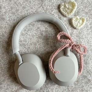 May include: Gray over-ear headphones with a pink crocheted bow and two white crocheted hearts. The headphones have a padded headband and earcups. The background is a floral patterned fabric.