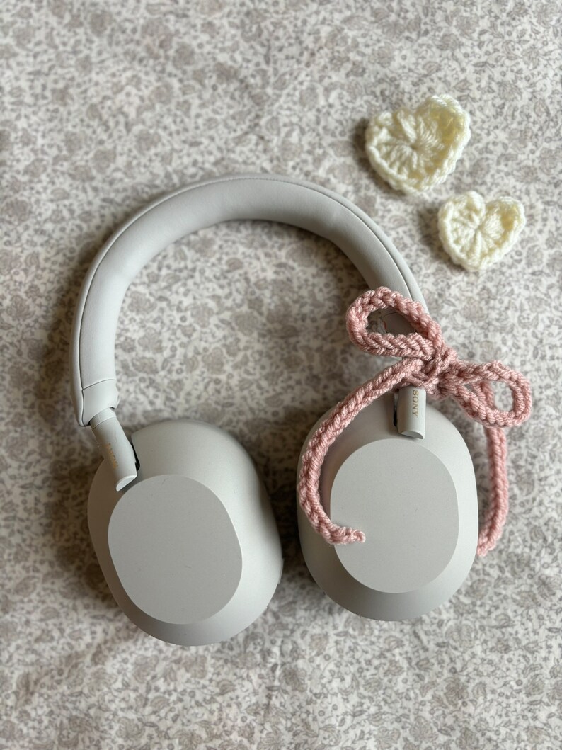 May include: Gray over-ear headphones with a pink crocheted bow and two white crocheted hearts. The headphones have a padded headband and earcups. The background is a floral patterned fabric.