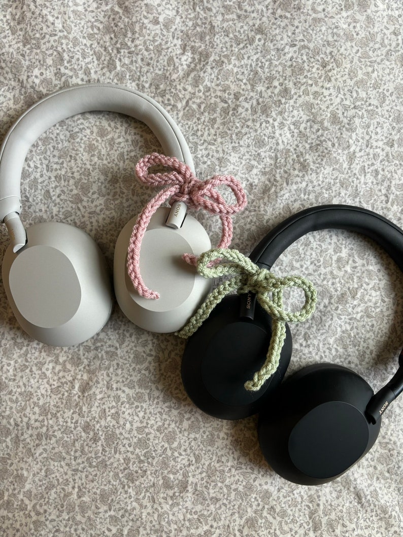 May include: Three pairs of Sony headphones in gray, black, and white, each adorned with a crocheted bow in pink and green. The headphones rest on a floral patterned fabric.