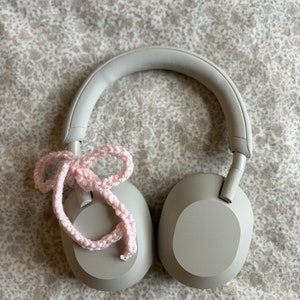 May include: Gray over-ear headphones with a light pink crocheted bow tied on the left earcup. The headphones are resting on a floral patterned surface. The earcups are oval-shaped.