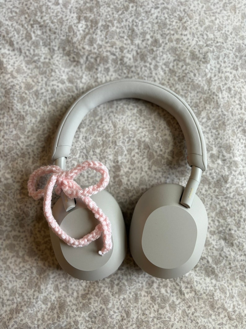 May include: Gray over-ear headphones with a light pink crocheted bow tied on the left earcup. The headphones are resting on a floral patterned surface. The earcups are oval-shaped.