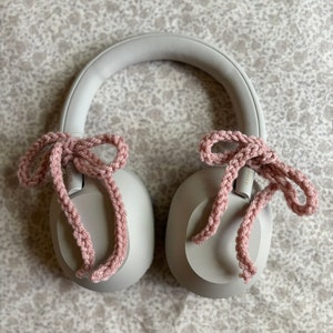 May include: A pair of gray headphones with pink crocheted bows attached to the earcups. The headphones are resting on a patterned surface. The bows are made of a soft, textured yarn.