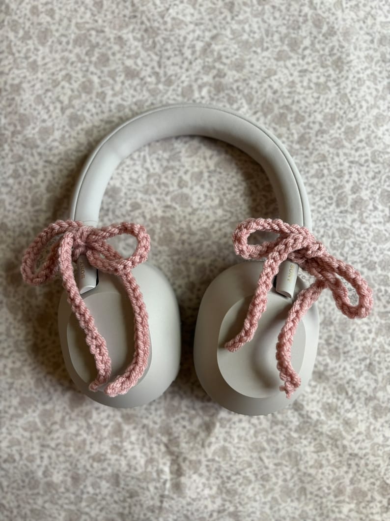 May include: A pair of gray headphones with pink crocheted bows attached to the earcups. The headphones are resting on a patterned surface. The bows are made of a soft, textured yarn.