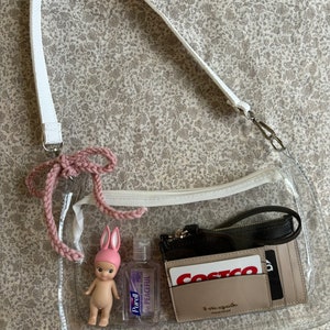 May include: A clear, rectangular crossbody bag with a white strap and a pink crocheted accent. Inside are a small doll, hand sanitizer, and a wallet with cards. The bag is designed to be see-through, showcasing the contents.