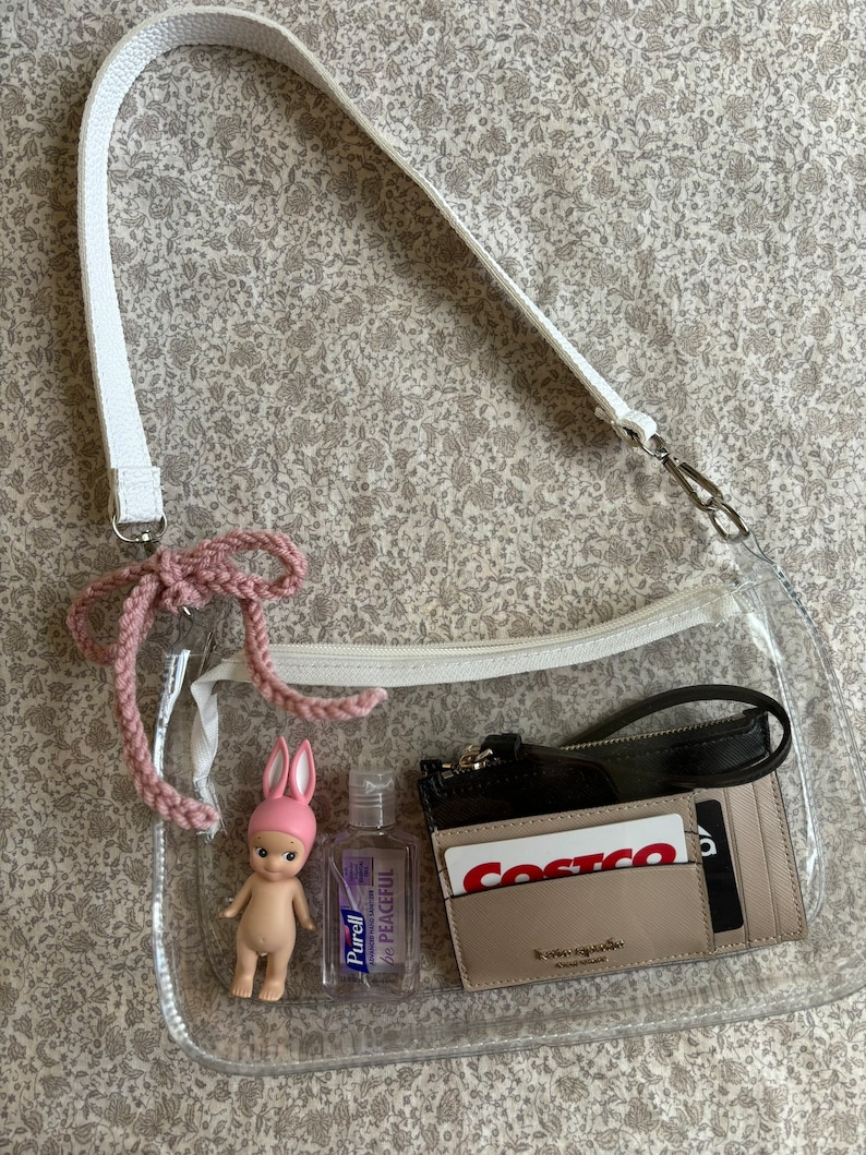 May include: A clear, rectangular crossbody bag with a white strap and a pink crocheted accent. Inside are a small doll, hand sanitizer, and a wallet with cards. The bag is designed to be see-through, showcasing the contents.