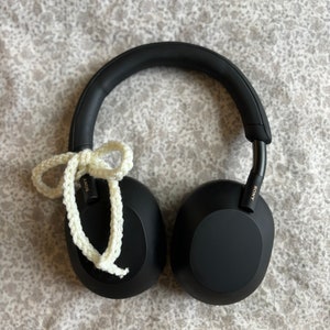 May include: Black over-ear headphones with a white crocheted cord tied in a bow. The headphones have a padded headband and earcups. The Sony logo is visible on the side of the headphones. The headphones are resting on a floral patterned surface.