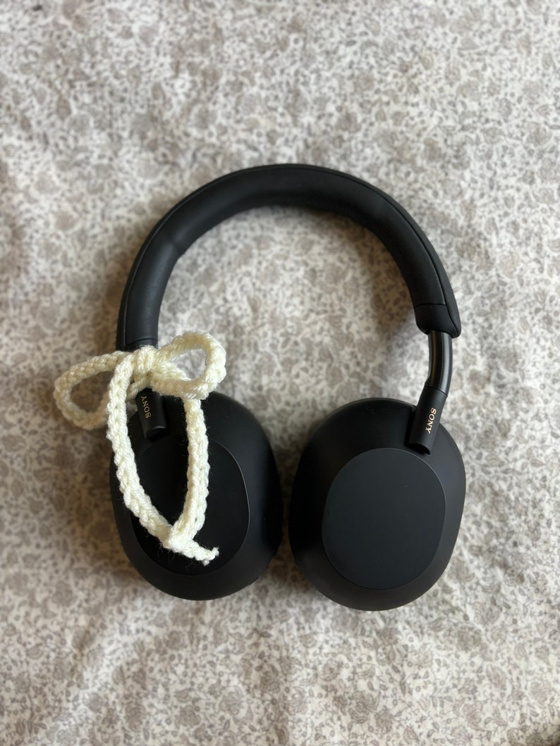 May include: Black over-ear headphones with a white crocheted cord tied in a bow. The headphones have a padded headband and earcups. The Sony logo is visible on the side of the headphones. The headphones are resting on a floral patterned surface.