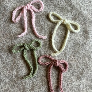 May include: Four handmade crochet bows in pastel colors: light pink, cream, sage green, and mauve. The bows are arranged on a floral patterned fabric. Each bow has a knotted center and long, dangling tails, showcasing a delicate, handcrafted aesthetic.