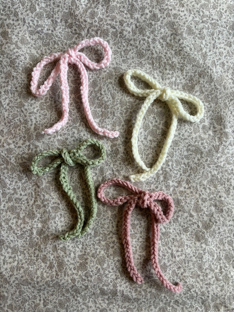 May include: Four handmade crochet bows in pastel colors: light pink, cream, sage green, and mauve. The bows are arranged on a floral patterned fabric. Each bow has a knotted center and long, dangling tails, showcasing a delicate, handcrafted aesthetic.