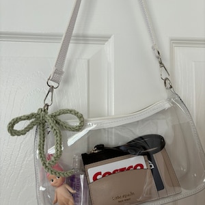 May include: A clear, rectangular shoulder bag with a white strap and zipper. Inside are a small doll, hand sanitizer, a Kate Spade cardholder, and a Costco card. A green crocheted bow hangs from the bag.