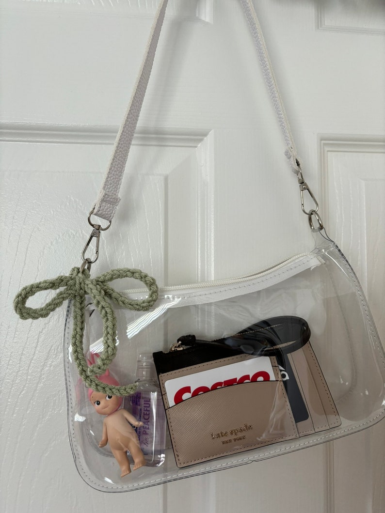 May include: A clear, rectangular shoulder bag with a white strap and zipper. Inside are a small doll, hand sanitizer, a Kate Spade cardholder, and a Costco card. A green crocheted bow hangs from the bag.