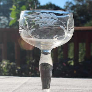 May include: A clear glass wine glass with a white etched floral design. The glass has a long stem and a wide bowl.