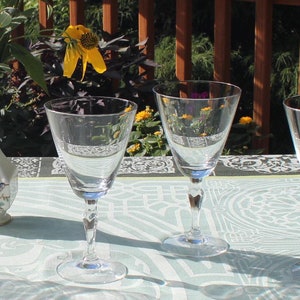 May include: Six clear glass wine glasses are arranged in a row on a white tablecloth with a green and white pattern. A vase with yellow and white flowers sits in the center of the table.